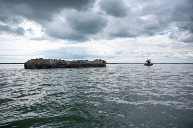 A tugboat pulls what looks like a bit of a rockly island across rippling waters.