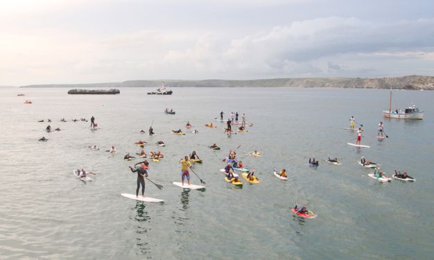 About forty people paddleboard in a body of water in front of a rocky island being pulled by a tugboat.
