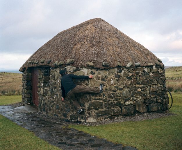 About a foot off the ground, a man hugs the corner of a thatched hut, hands and feet gripping the stones.