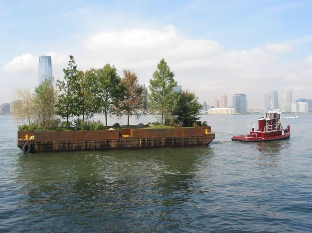 Against the New York City skyline, a tugboat pulls a barge planted with trees, plants, and grass along a river.
