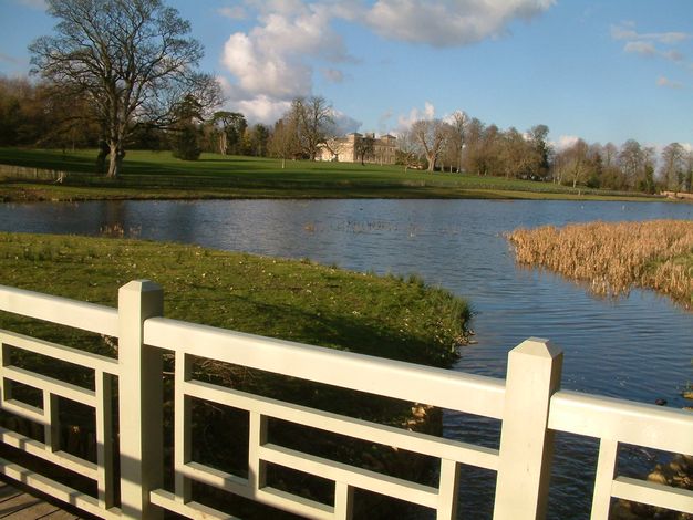 In low, sunset light, a lake with reeds growing at its centre leads back to a smooth green lawn and, in the distance, a stone manor.