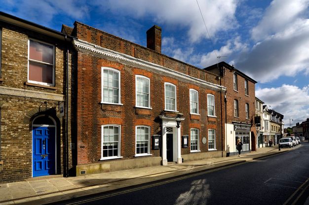 Architecture. A two-storey red brick building has a central door surrounded by white moulding. The four windows along the lower level and five above have slightly curved tops. White clouds skid towards us in a radiantly blue sky above.